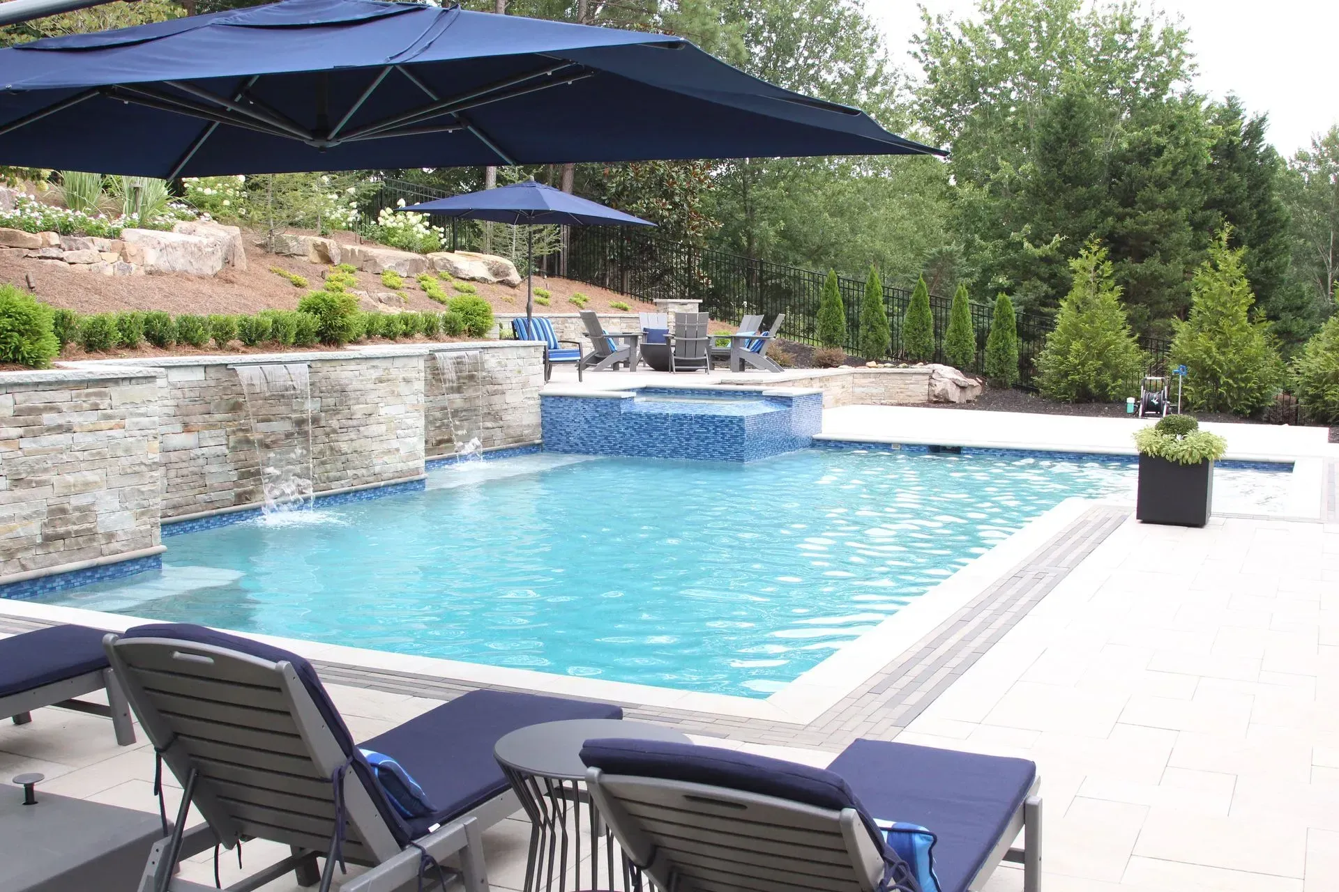 Swimming pool with blue umbrellas, lounge chairs, waterfall, and greenery.