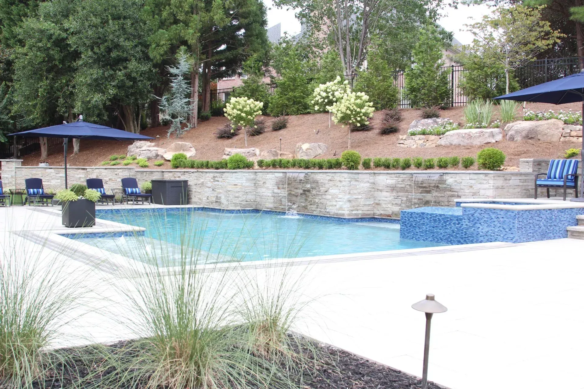Swimming pool with blue umbrellas, surrounded by landscaping, retaining wall, and trees.
