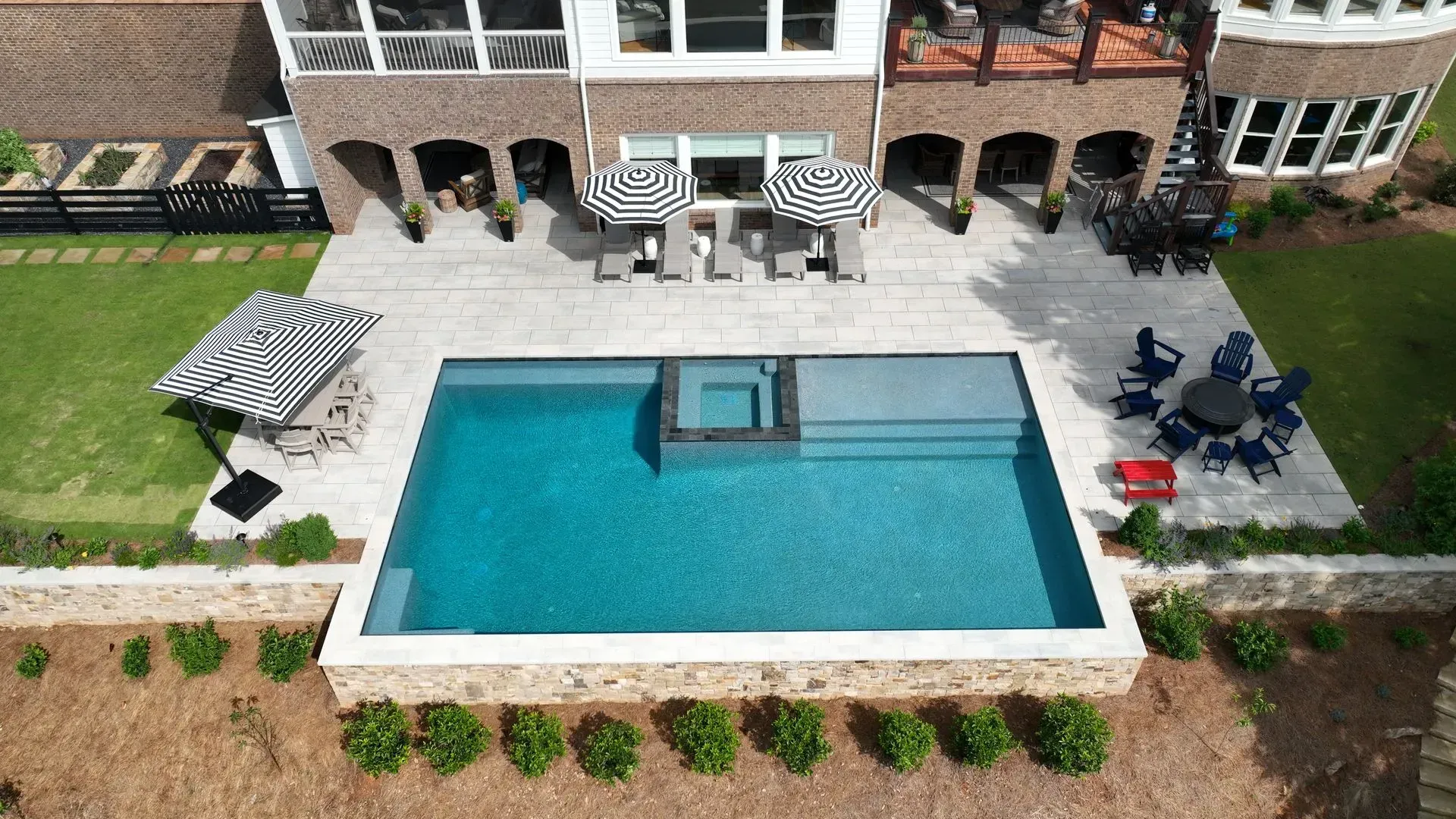 Aerial view of a rectangular pool with a spa. Beige patio, outdoor furniture, and a multi-story brick house in the background.
