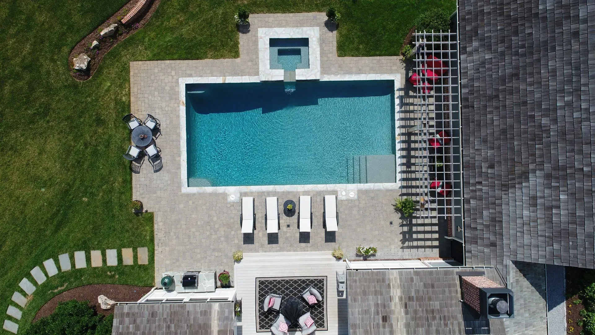 Overhead view of a rectangular pool with a hot tub, surrounded by a patio and a grassy lawn, near a shingled roof.