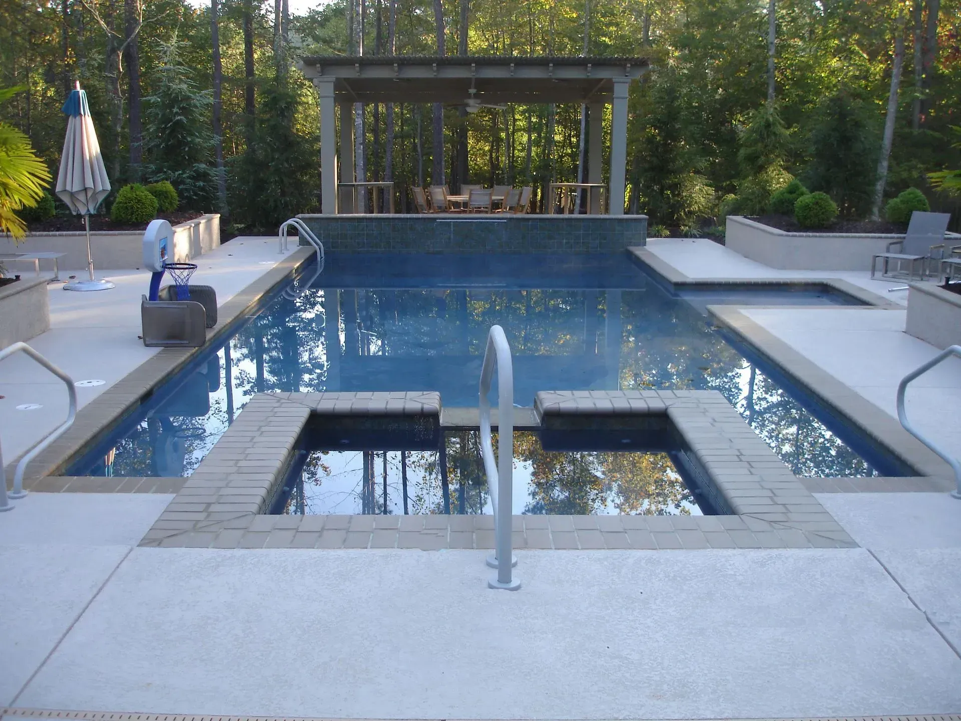 A luxurious backyard pool with a spa, patio, and dining area under a wooden pergola.
