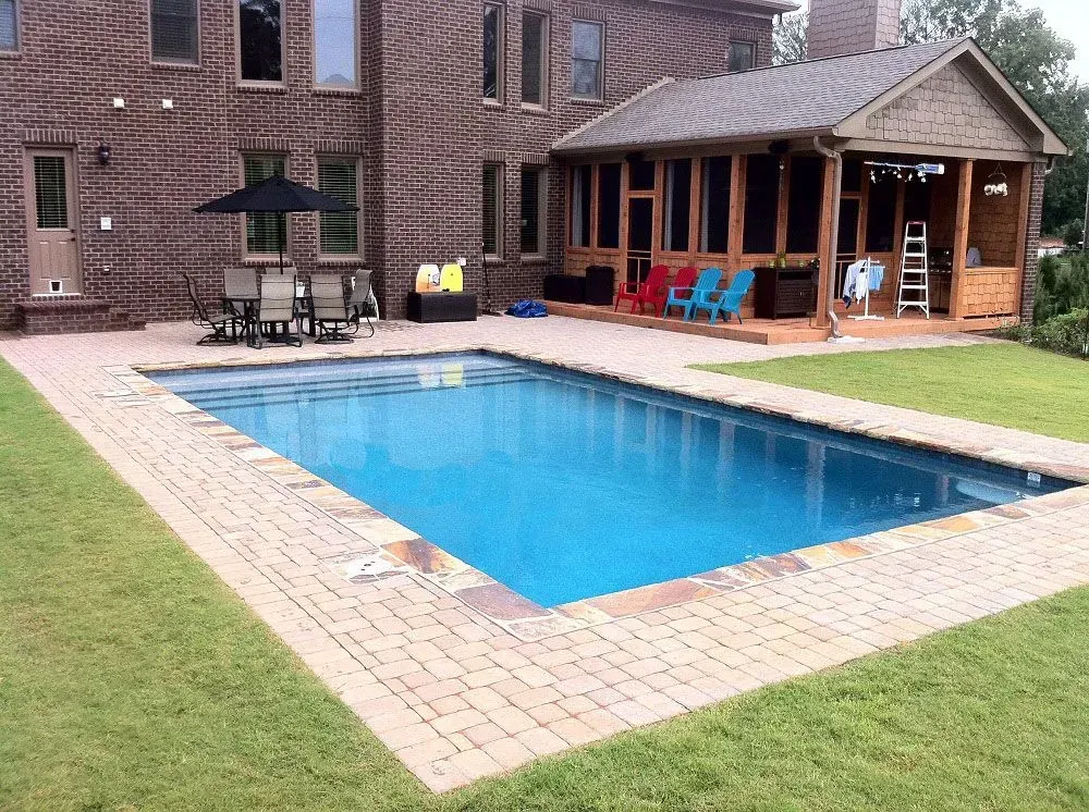 Rectangular pool with brick patio, surrounded by green grass, next to a brick house and screened-in porch.