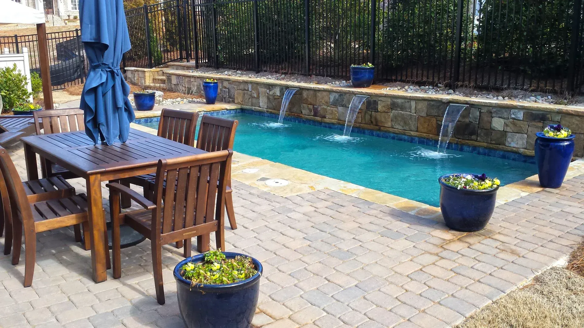 Outdoor pool area with a wooden table and chairs, blue umbrella, water fountains, and potted plants.