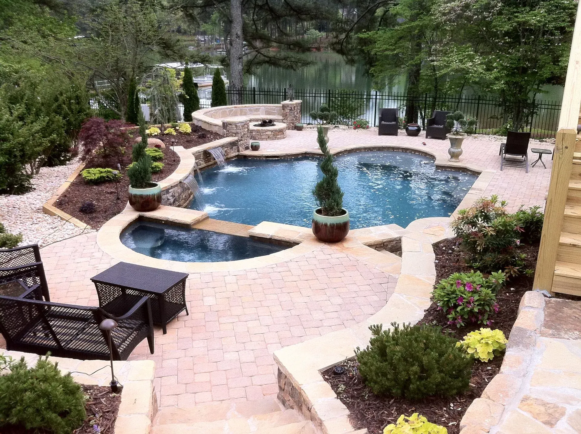 A backyard pool with waterfall, patio, and lounge seating; surrounded by landscaping, overlooking a lake.