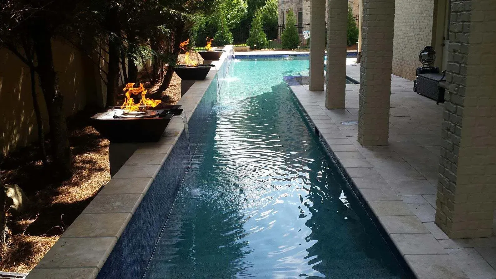 Long, narrow swimming pool with fire features, pillars, and landscaping.
