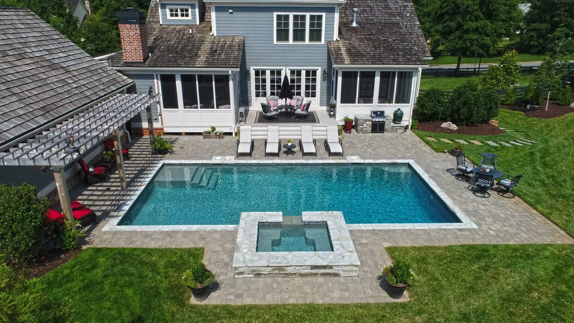 Backyard with blue swimming pool and spa, surrounded by stone patio, lush green grass, and a light blue house.
