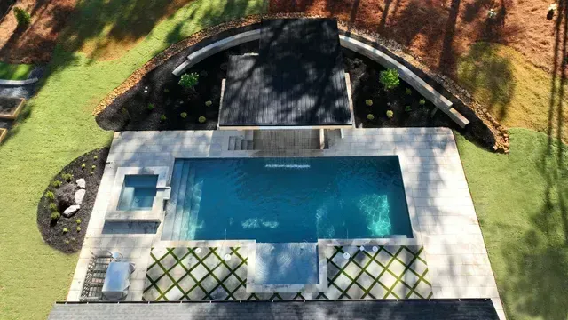 Aerial view of a rectangular pool with a hot tub, surrounded by a concrete patio and a curved, landscaped area.
