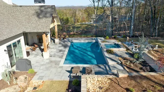 Backyard with rectangular pool and spa, patio, and landscaping, adjacent to a home with a gray roof and forested backdrop.