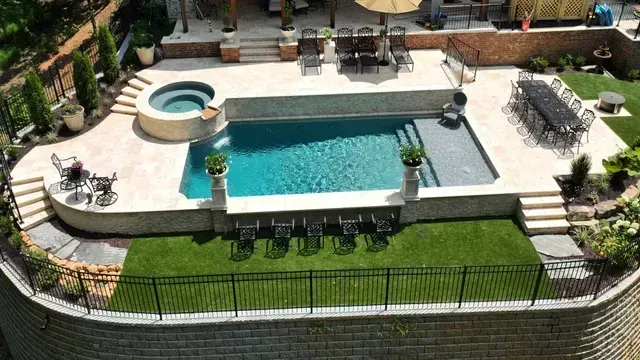 Aerial view of a backyard with a pool, hot tub, and patio, featuring landscaping with green grass and a black fence.
