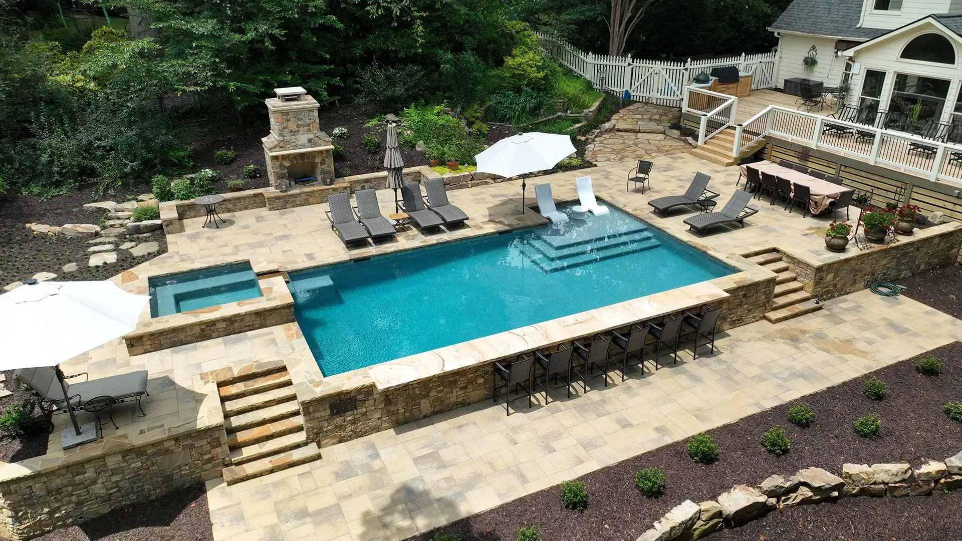 Aerial view of a rectangular turquoise pool with a spa, lounge chairs, and an outdoor dining area. Stone walls and a white house.