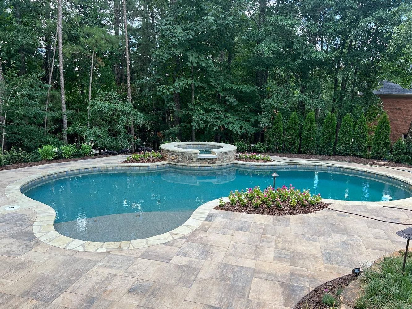 A kidney-shaped swimming pool with a connected hot tub, surrounded by stone patio and landscaping, set in a backyard.