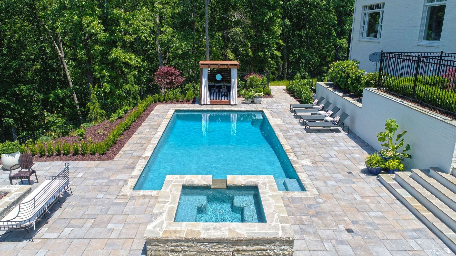 Pool with hot tub, stone patio, lounge chairs, and cabana; trees in background.