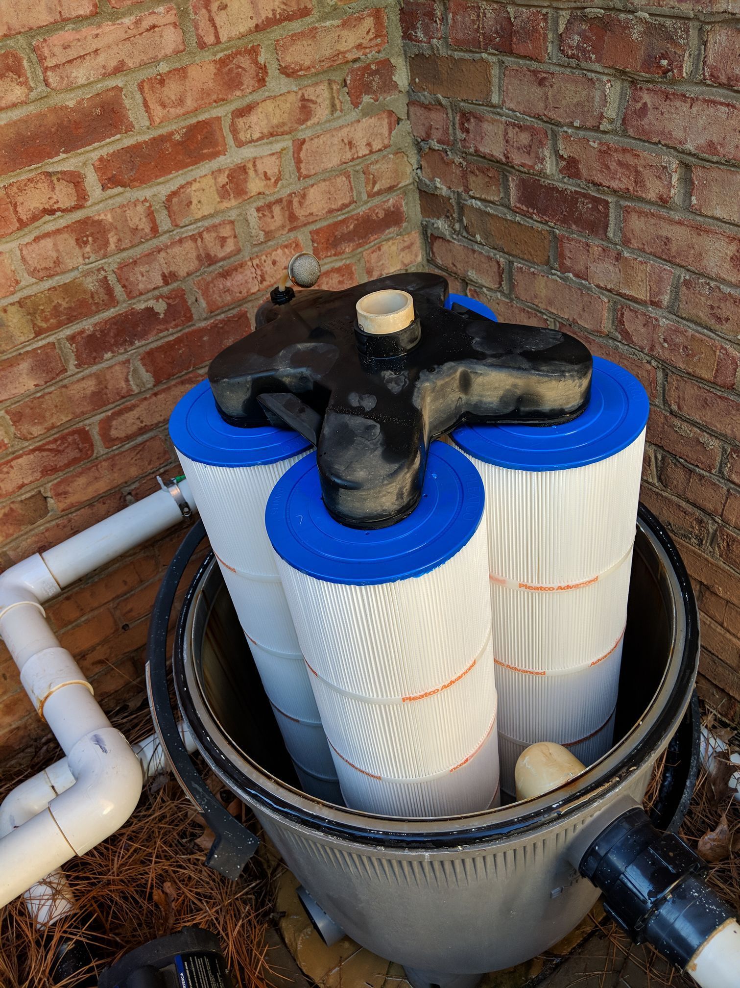 Pool filter system with three cylindrical filter cartridges inside a plastic housing, against a brick wall.