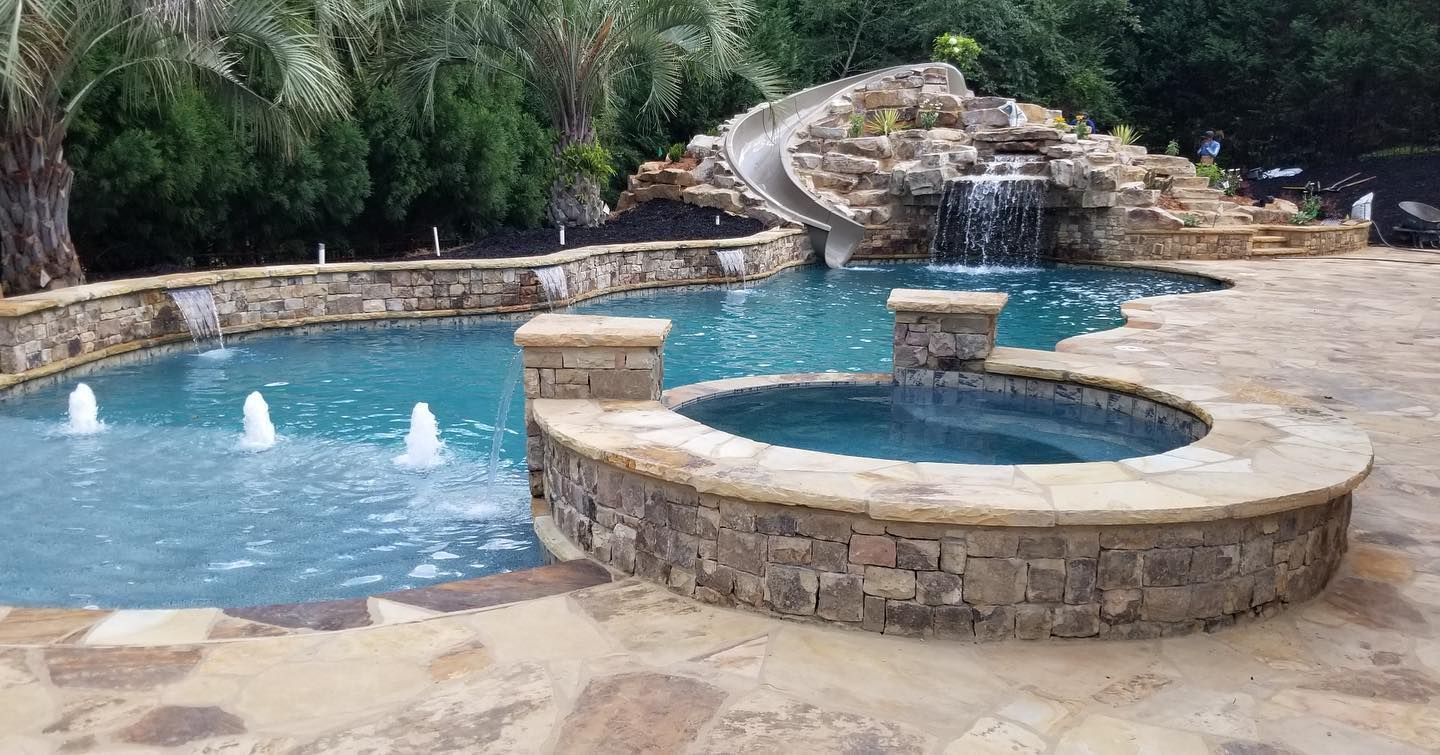 Luxury pool with waterfall, slide, and hot tub surrounded by stone and greenery.