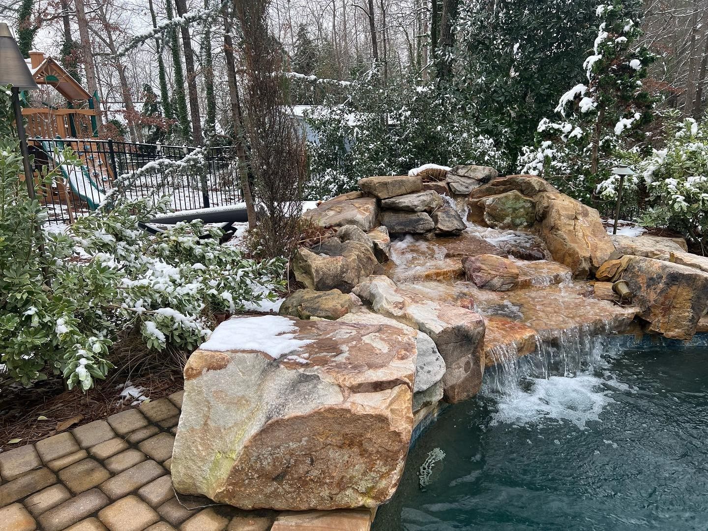Snowy scene of a waterfall cascading into a pool, with rocks, landscaping, and a wooden play structure in the background.