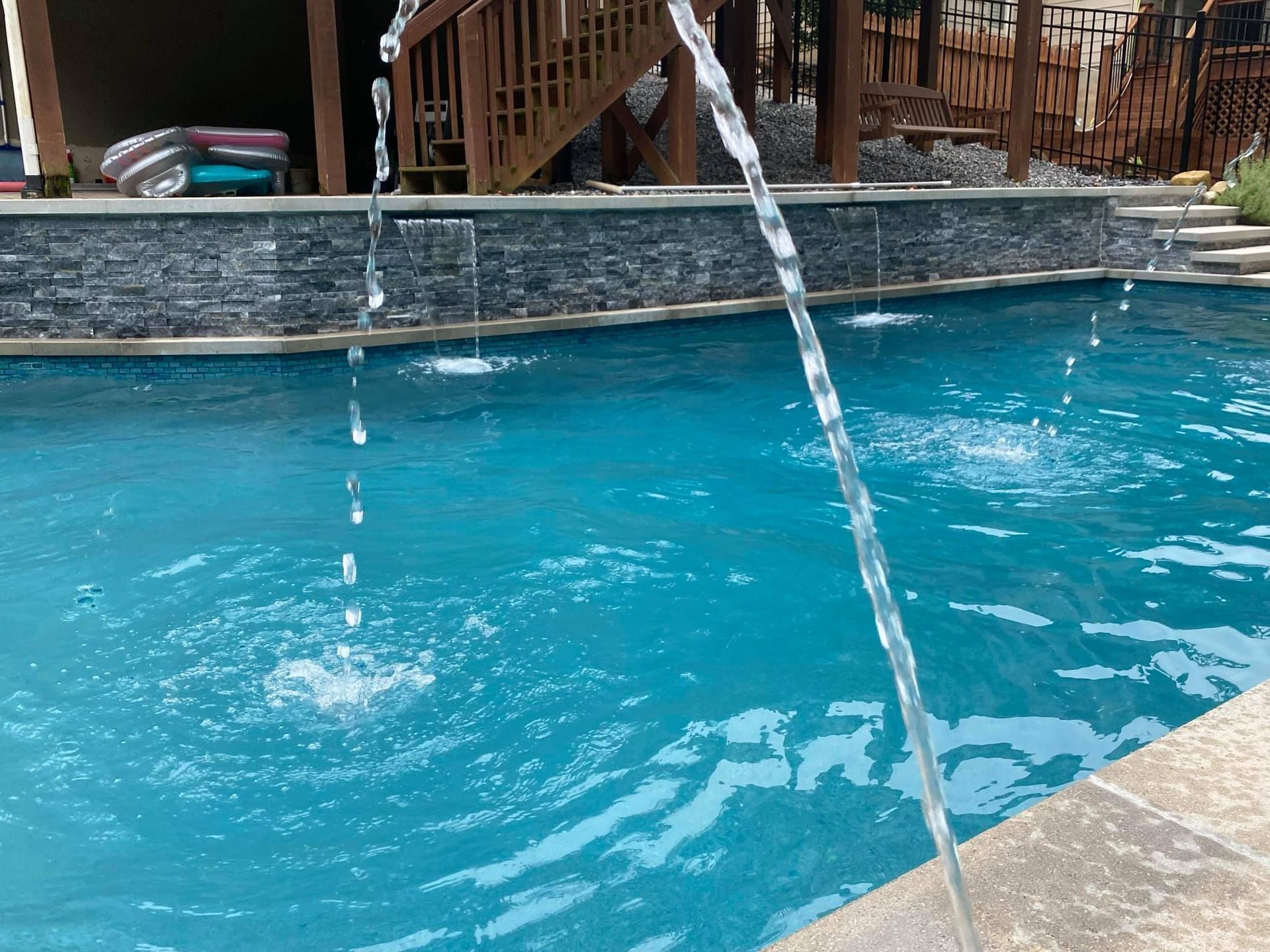 Pool with cascading water features; stone wall backdrop; sunny outdoor setting.