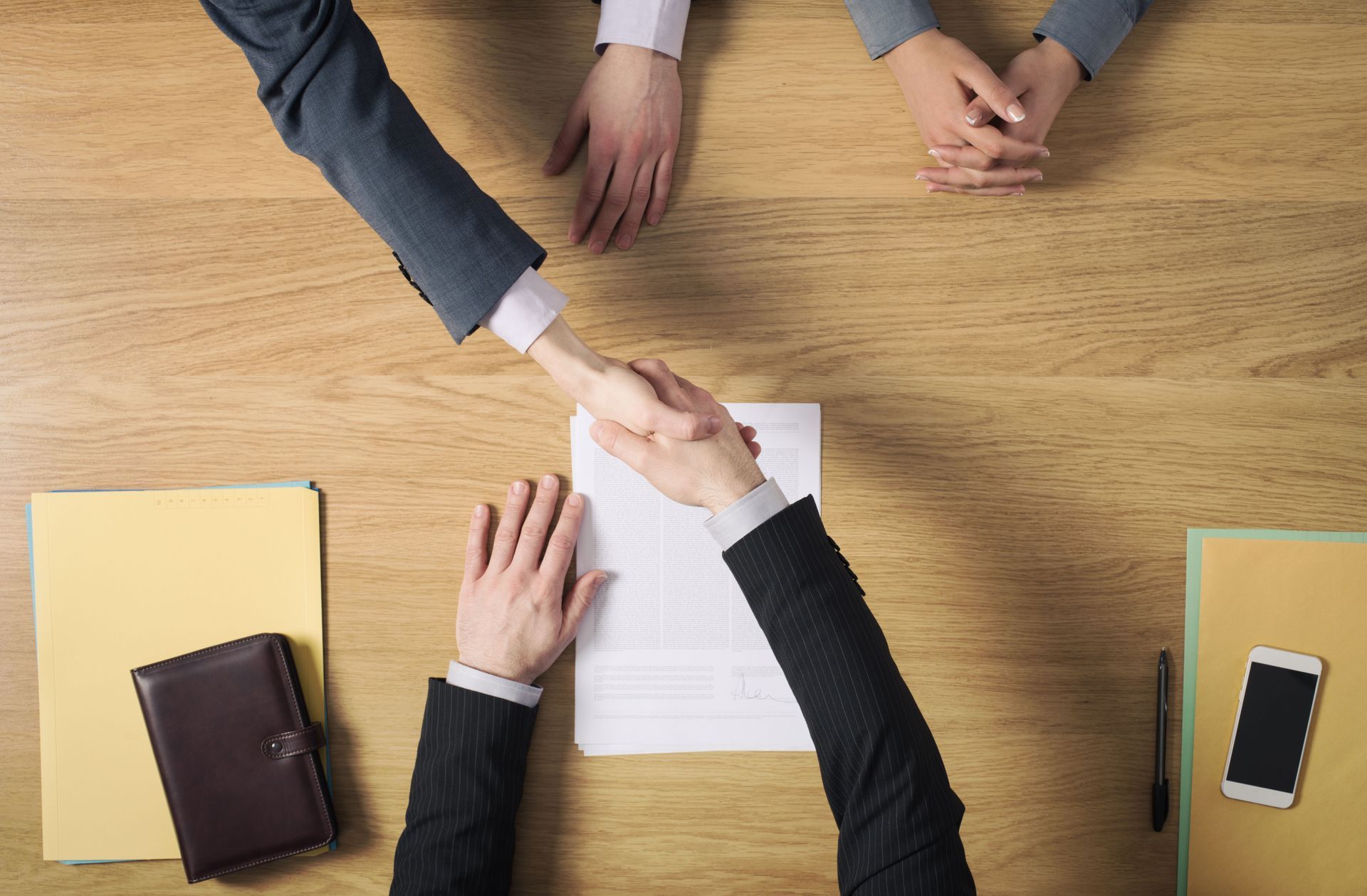 Business meeting handshake over a wooden table with documents, notebooks, and a phone.