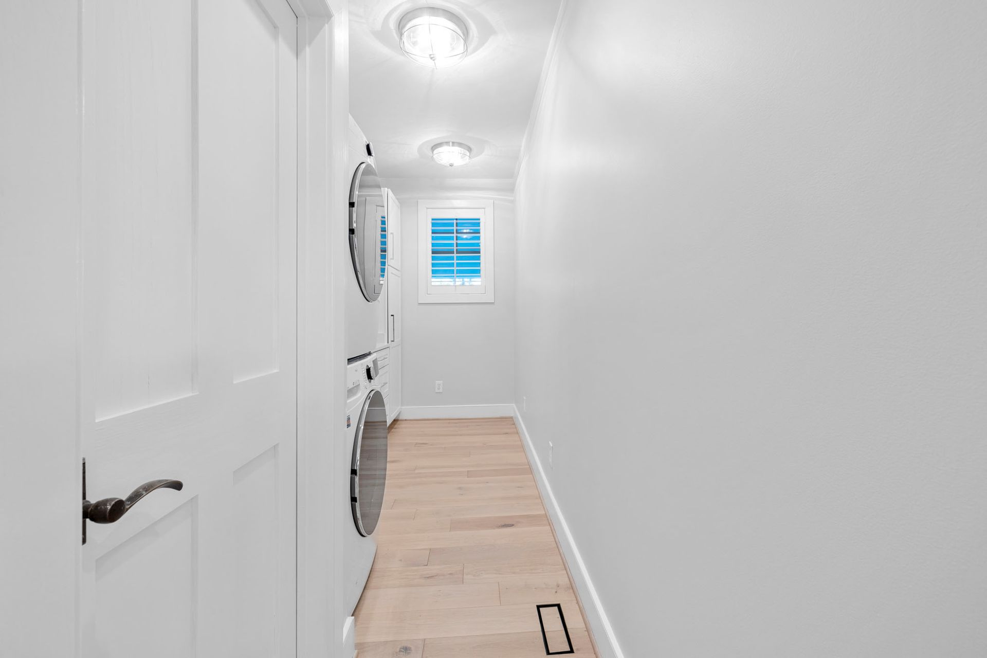 Narrow white laundry hallway with stacked washer and dryer, light wood floor, and window at the end.