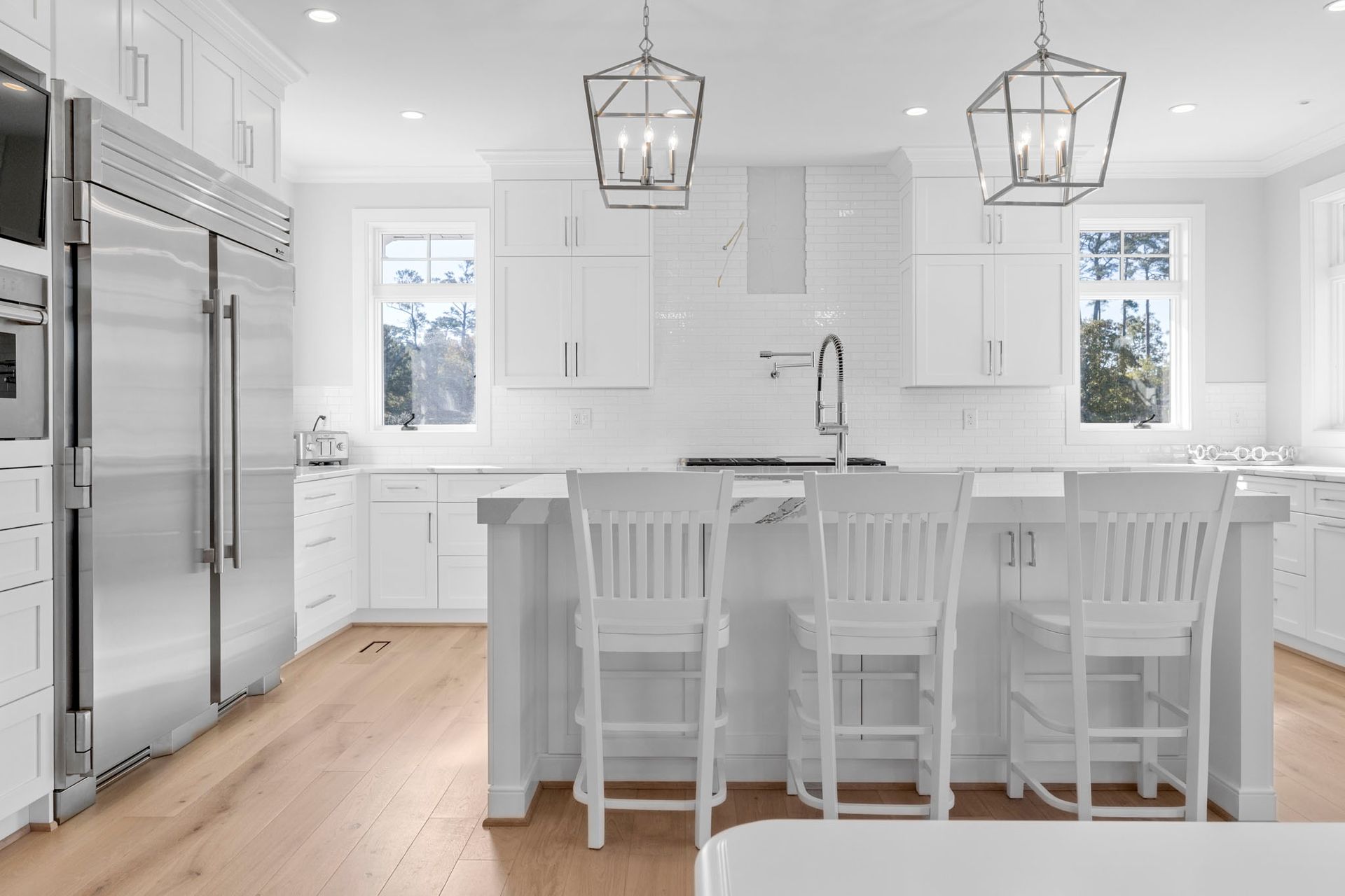 Bright white kitchen with island, bar stools, stainless fridge, and pendant lights above light wood floor.