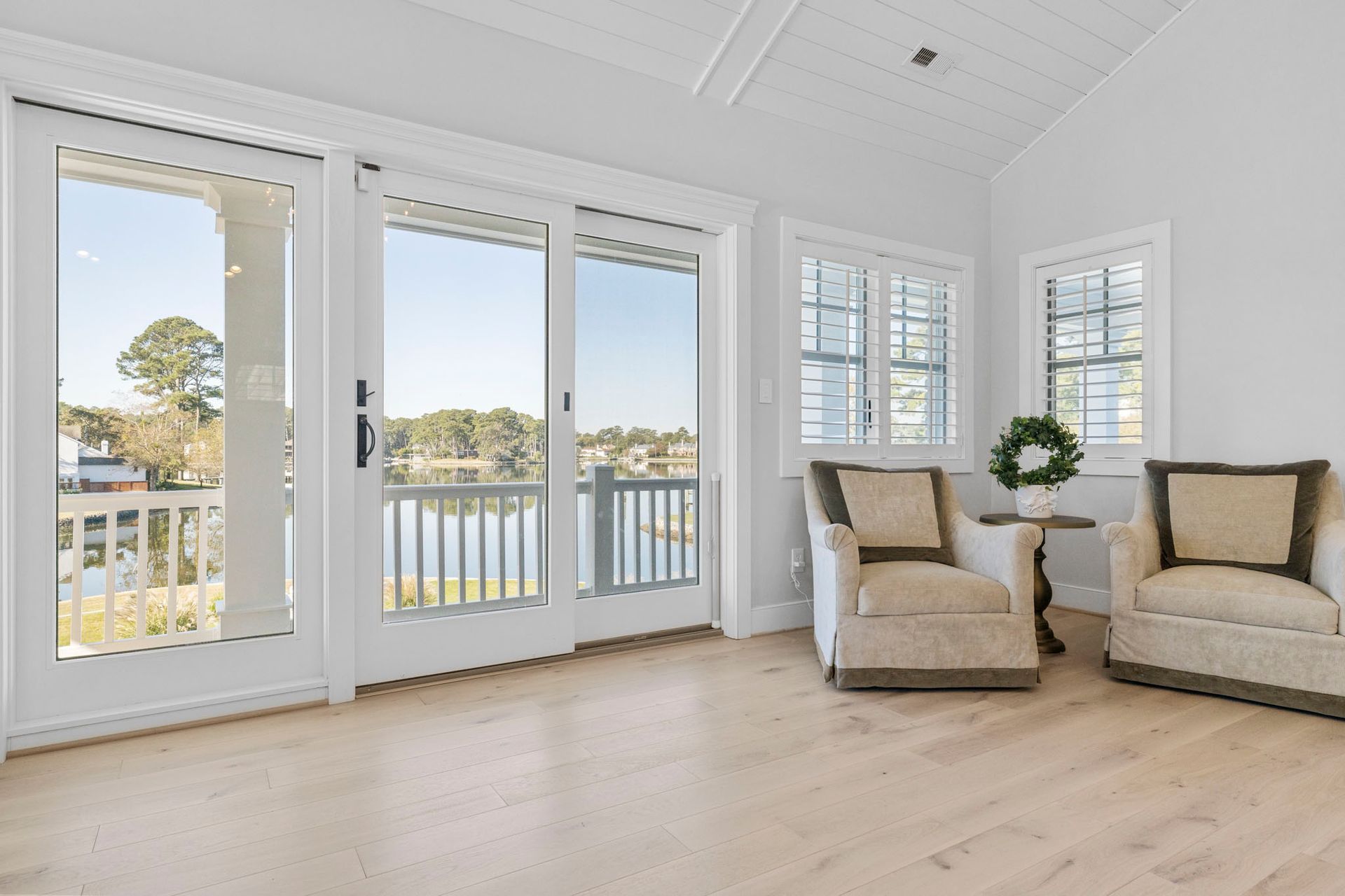 Bright sunroom with white walls, French doors, and two beige armchairs overlooking water and trees.