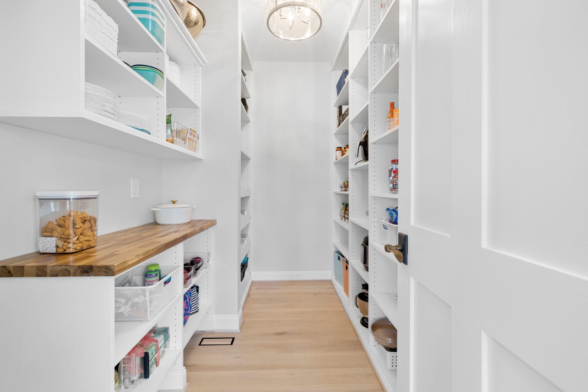 Bright white walk-in pantry with open shelves, wooden counters, and jars along a narrow hallway.