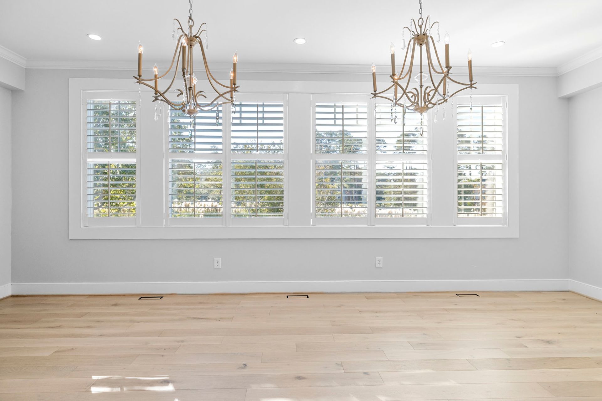 Bright empty dining room with large windows, white shutters, gold chandeliers, and light wood floors
