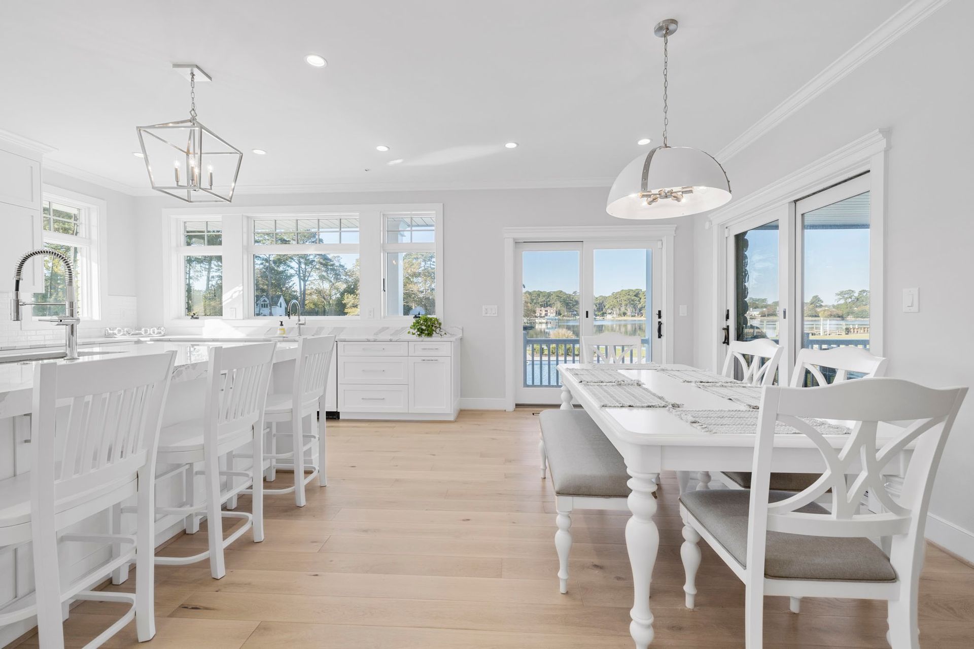 Bright white dining room with long table, chairs, pendant lights, and windows overlooking the water.