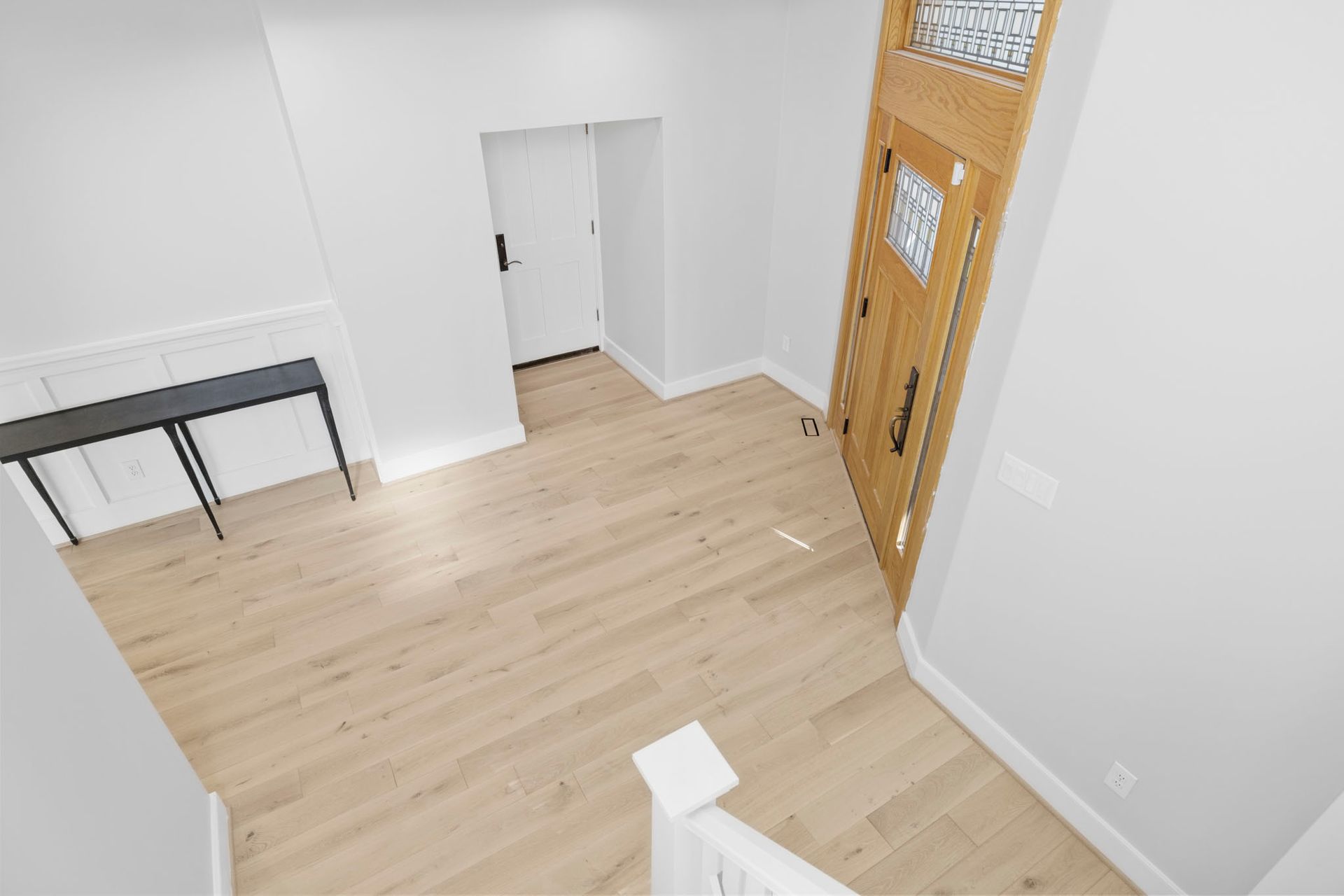 Empty room with light wood floor, white walls, and a wooden door near a small table.