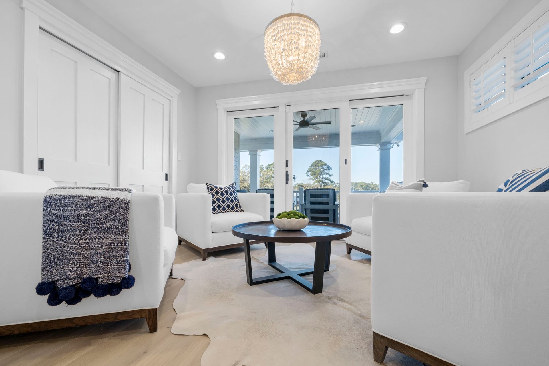 Bright living room with white sofas, blue accents, round coffee table, and sliding glass doors to a patio.