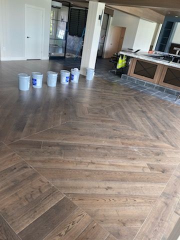 Empty room with herringbone wood floor, paint buckets lined up, and kitchen cabinets in the background