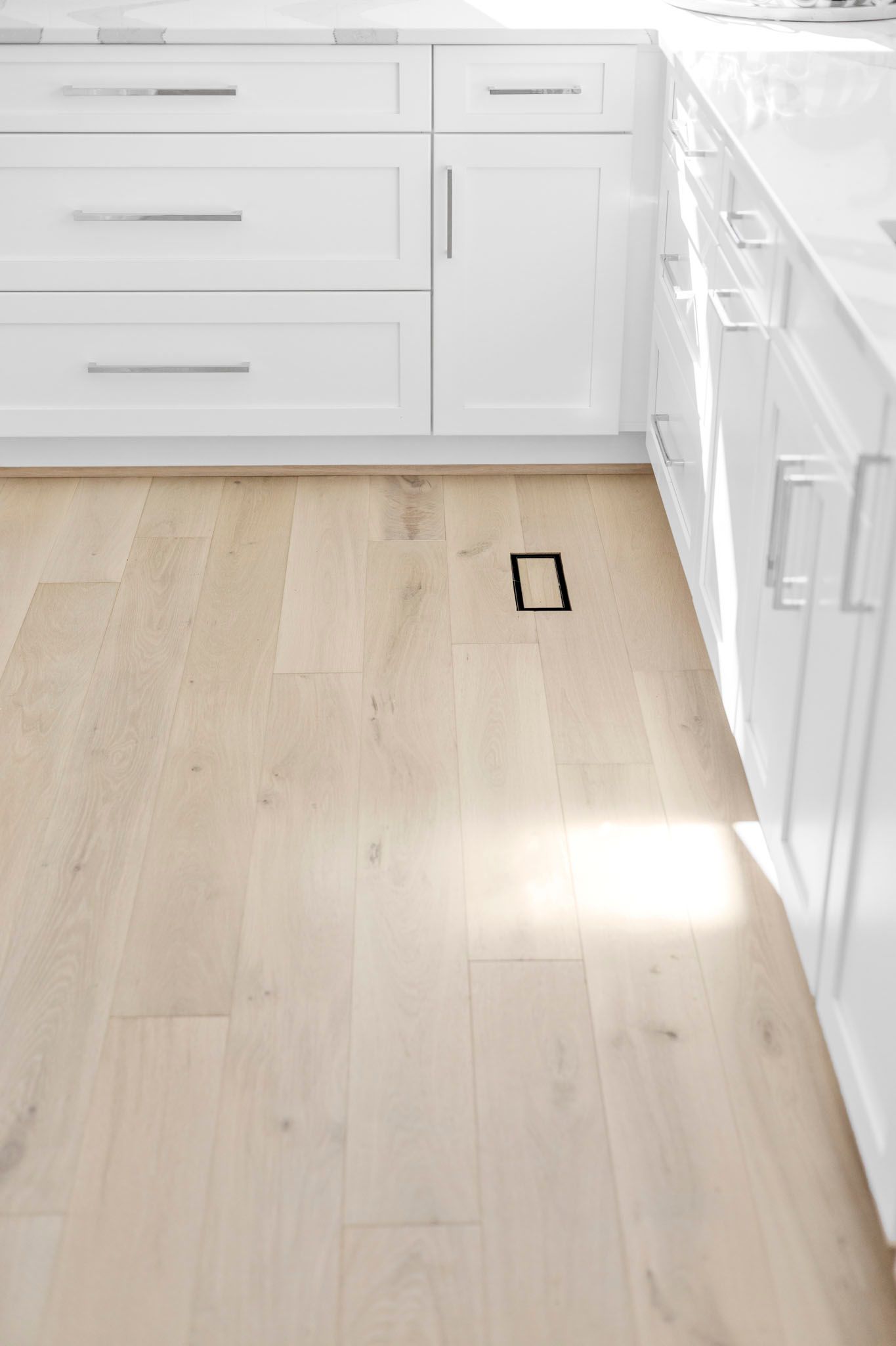 Bright kitchen with white cabinets, pale wood floor, and a small floor vent or opening near the center.