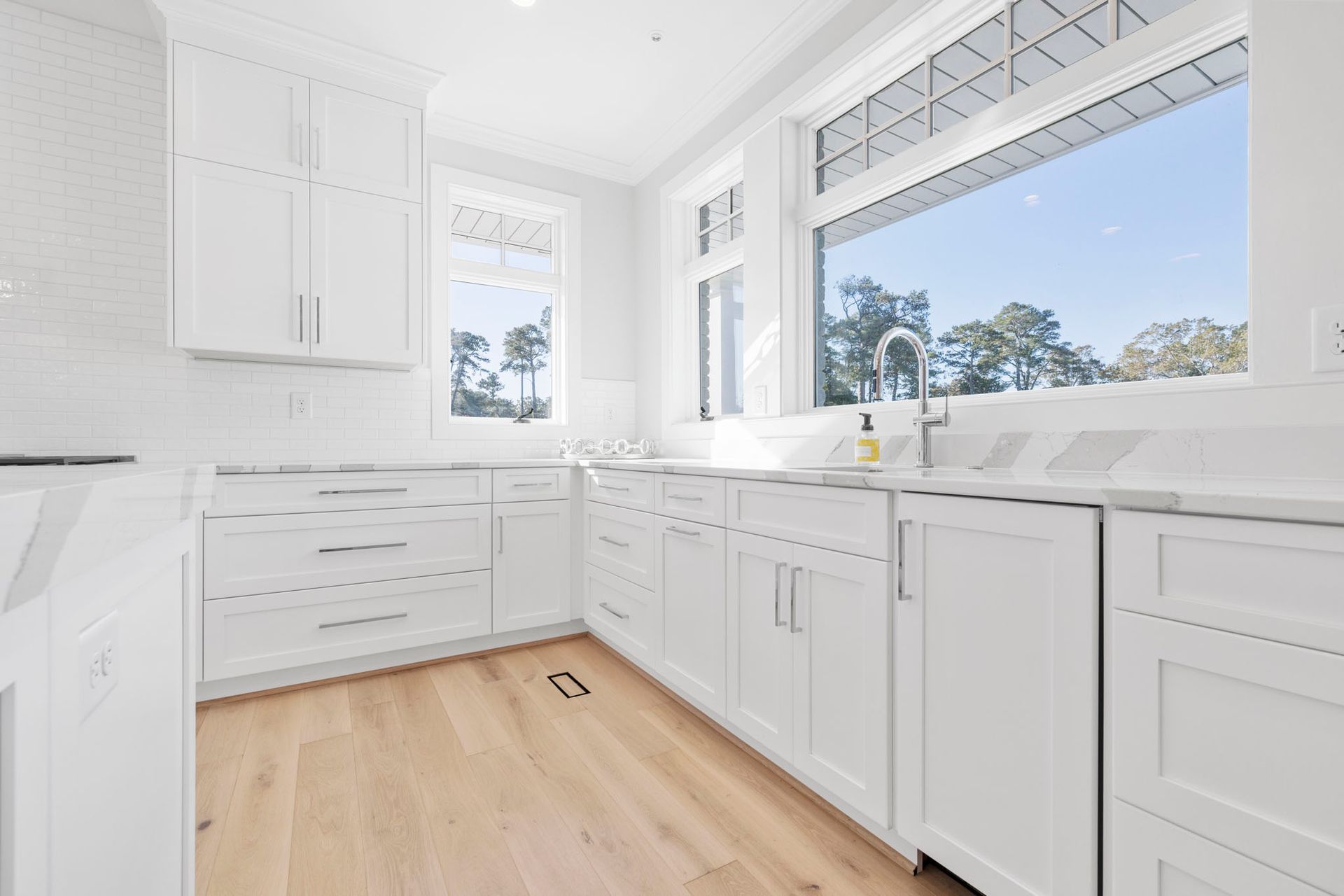 Bright white kitchen with wood floors, large windows, and a sink under sunny daylight