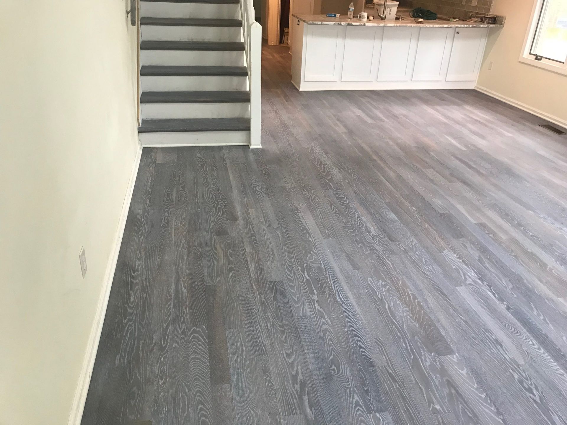 Empty living room with gray flooring, staircase, and kitchen island in the background