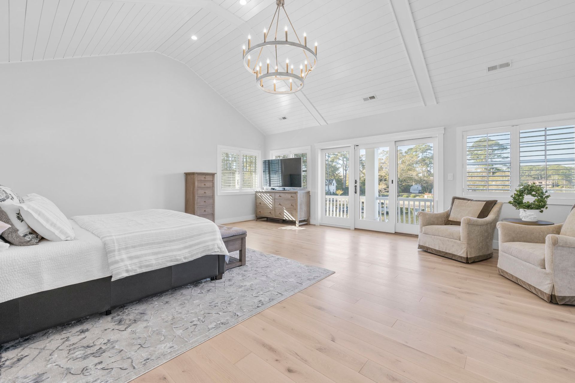 Bright bedroom with vaulted ceiling, chandelier, bed, armchairs, and large windows overlooking greenery