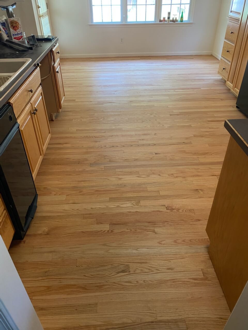 Empty kitchen with light wood floors, cabinets, and a window at the far end.