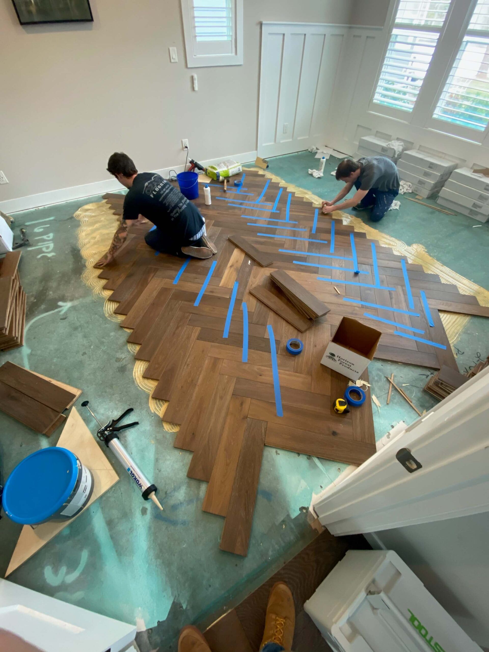 Two people laying a herringbone wood floor in a bright room, with tools and blue tape visible.