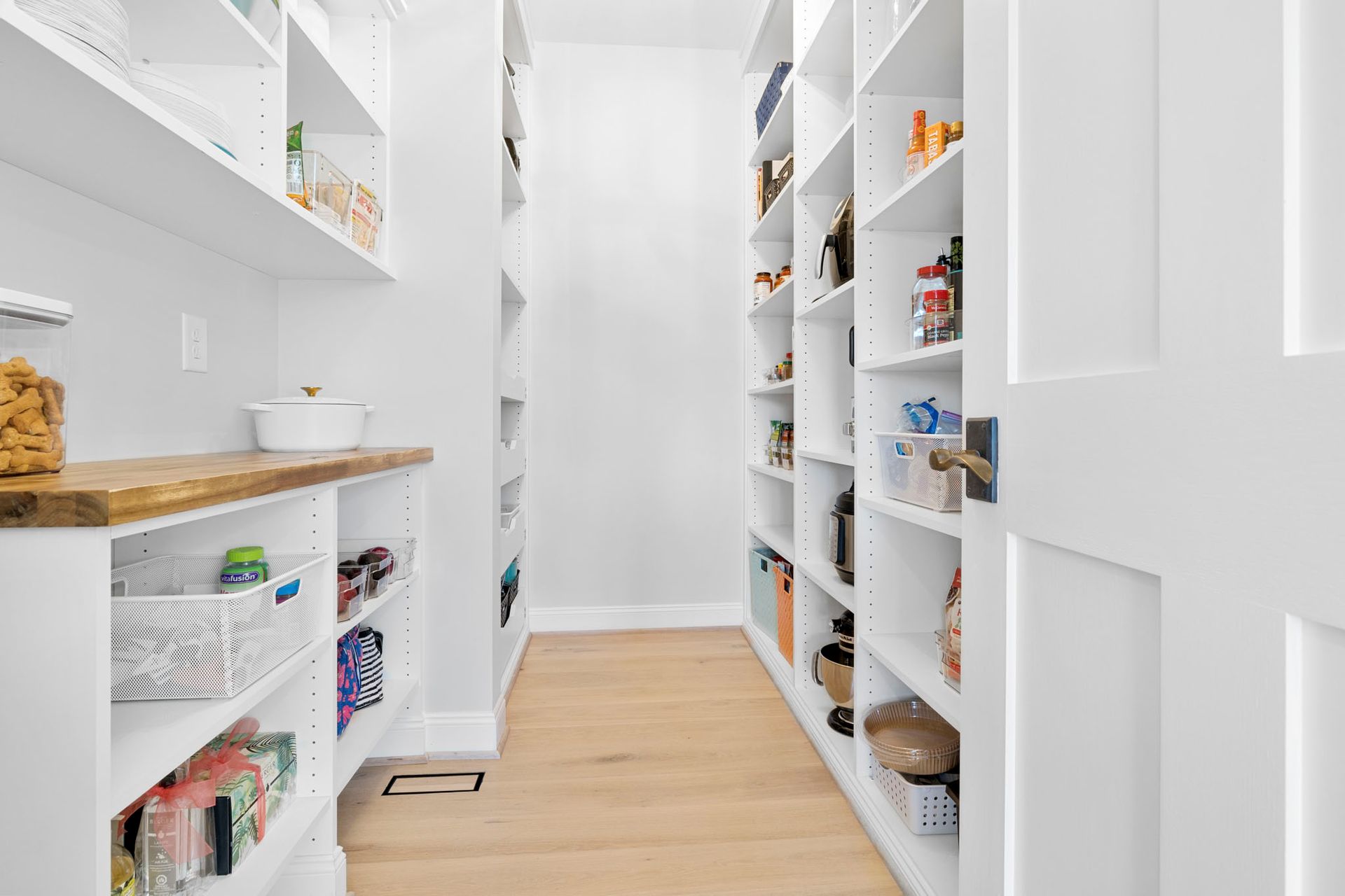 Bright white pantry with open shelves, wooden countertops, and wicker baskets along a narrow hallway