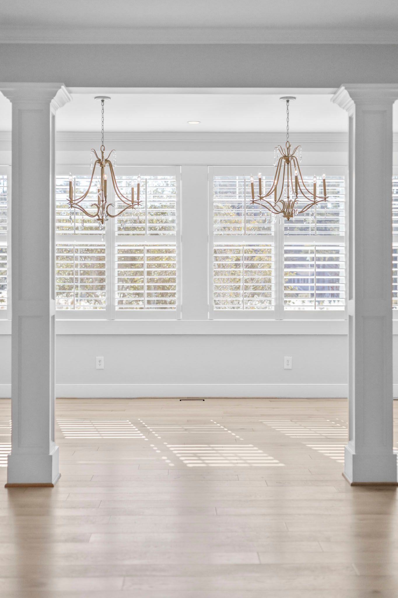 Bright empty living room with hardwood floors, large windows, and two gold chandeliers
