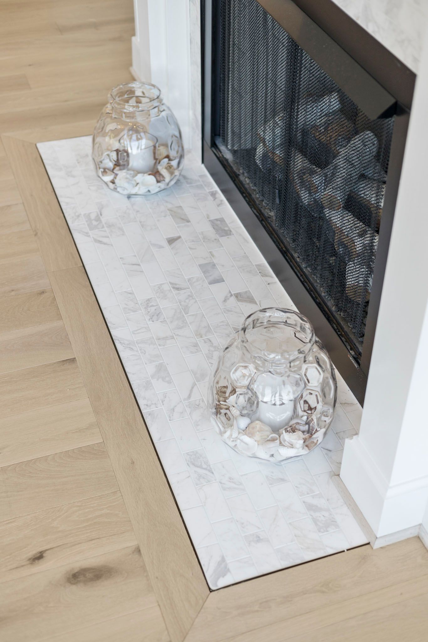 White faux-fur mat with two clear glass candle holders beside a black fireplace on a light wood floor