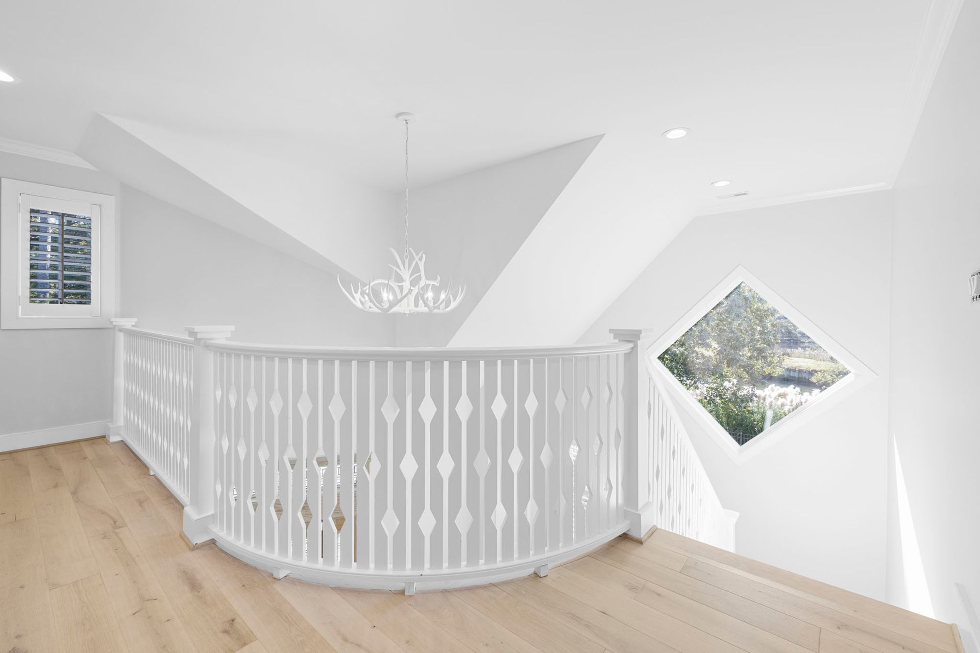 Bright white upstairs landing with curved railing, chandelier, wood floor, and geometric window.