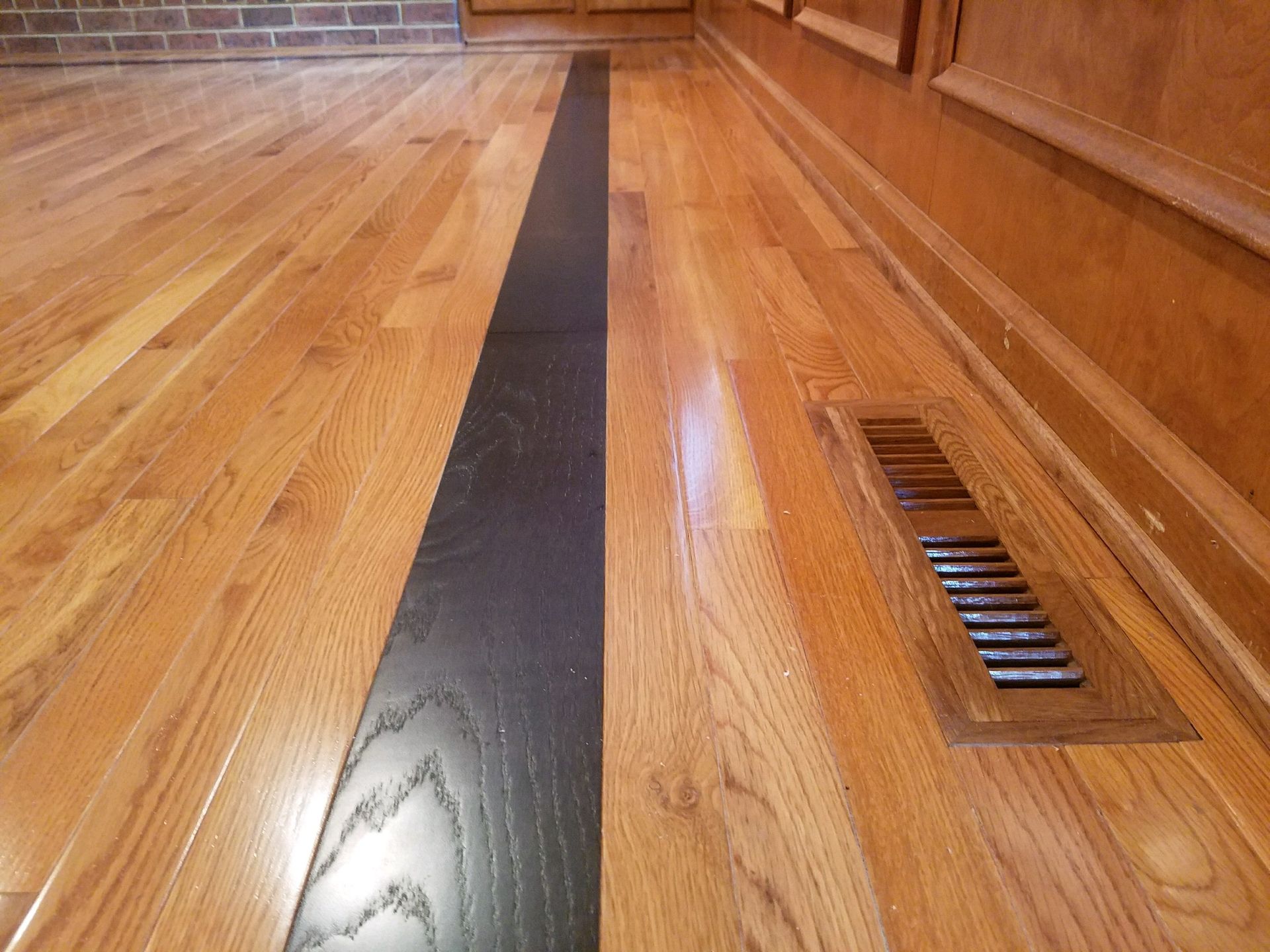 Wooden floor with a long black strip and a floor vent beside a wood wall panel