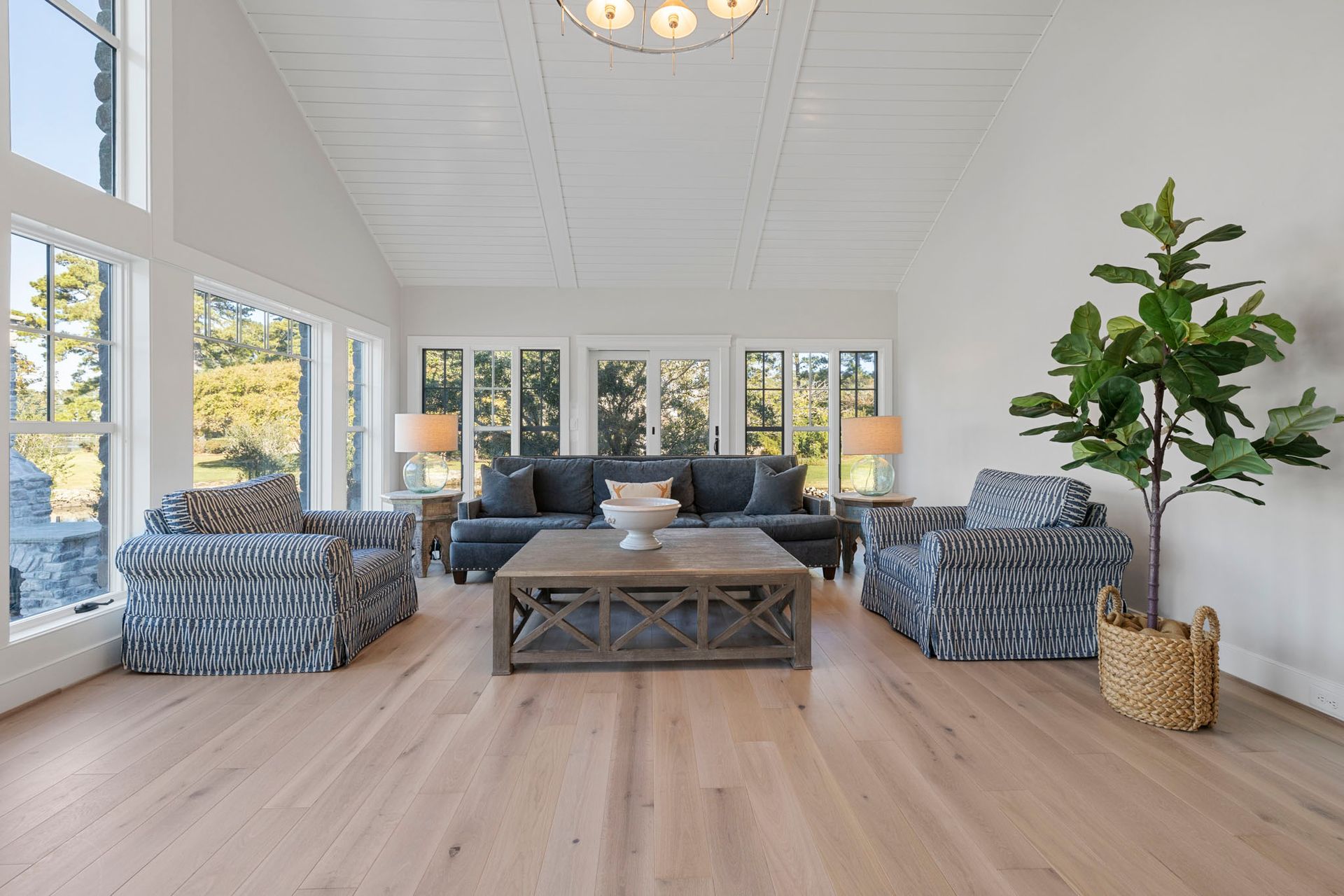 Bright sunroom with large windows, gray sofa, patterned chairs, coffee table, and a tall potted plant