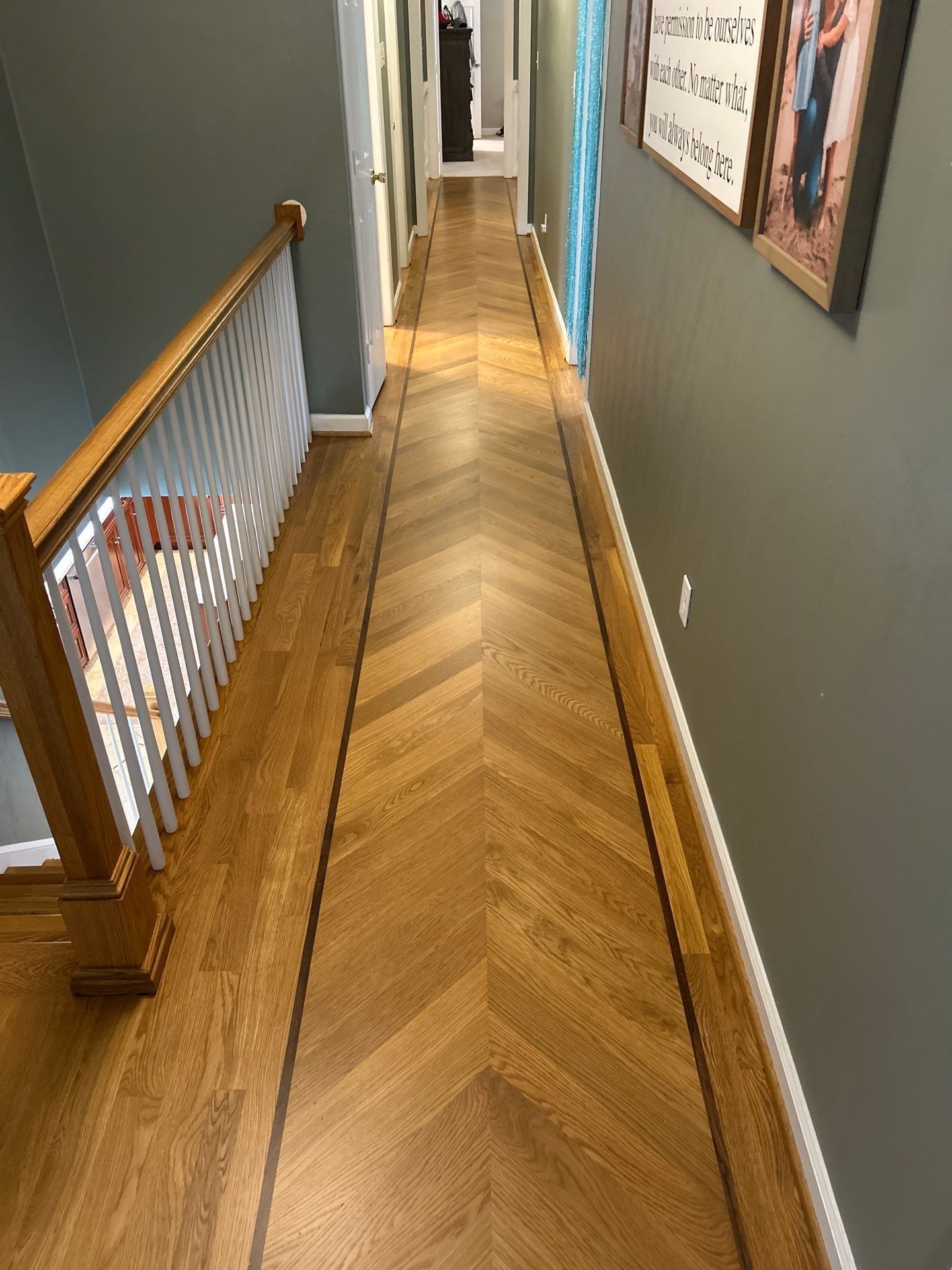 Narrow upstairs hallway with herringbone wood floor, gray walls, and framed art along the right side.