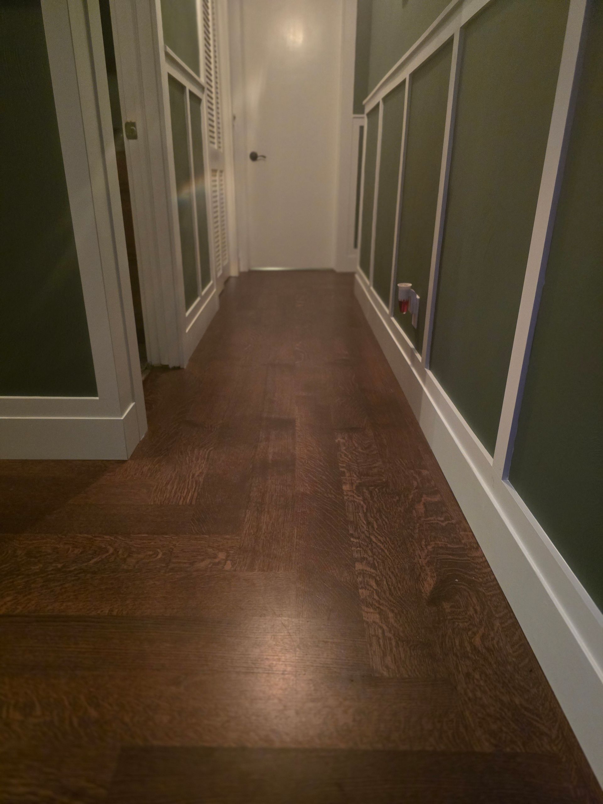 Narrow hallway with dark wood floors, green walls, and white trim, leading to a closed door.
