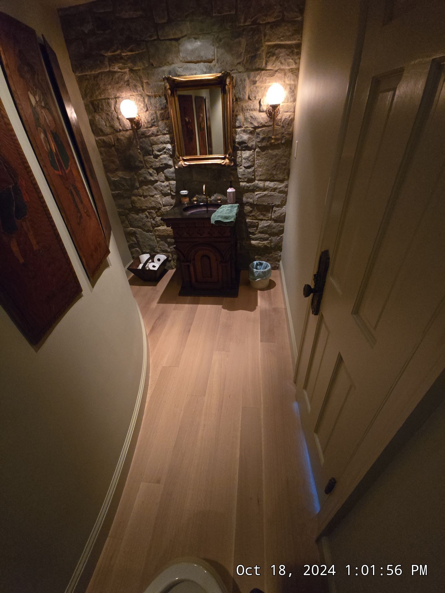 Dim hallway leading to a stone alcove with a mirror, cabinet, wall sconces, and a trash can.