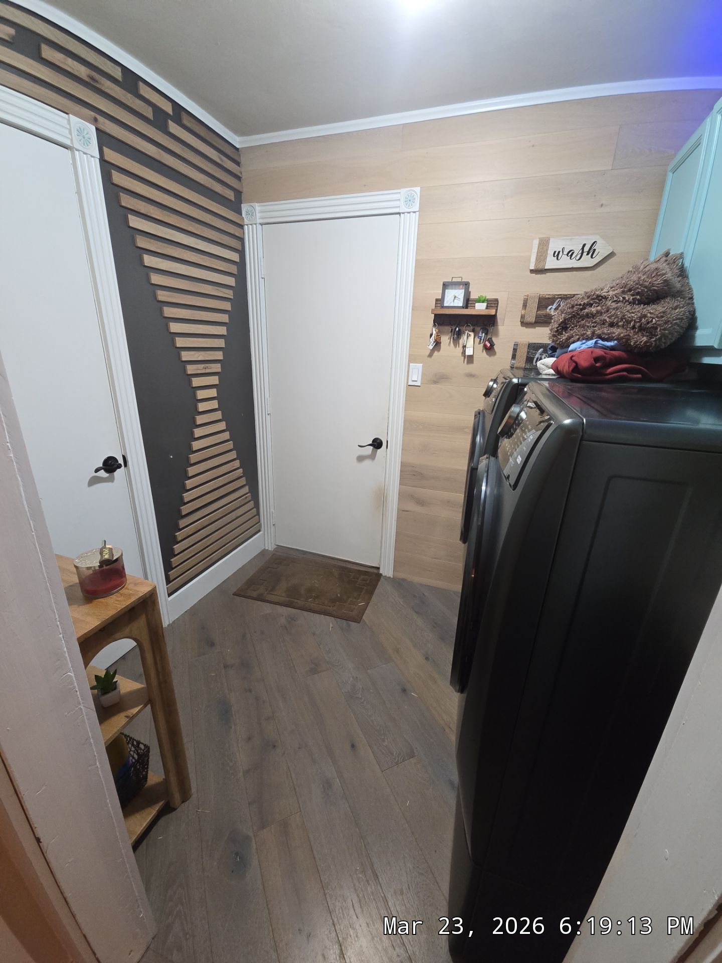 Small cluttered room with wood floors, white door, striped wall, and stacked bins beside a black appliance.