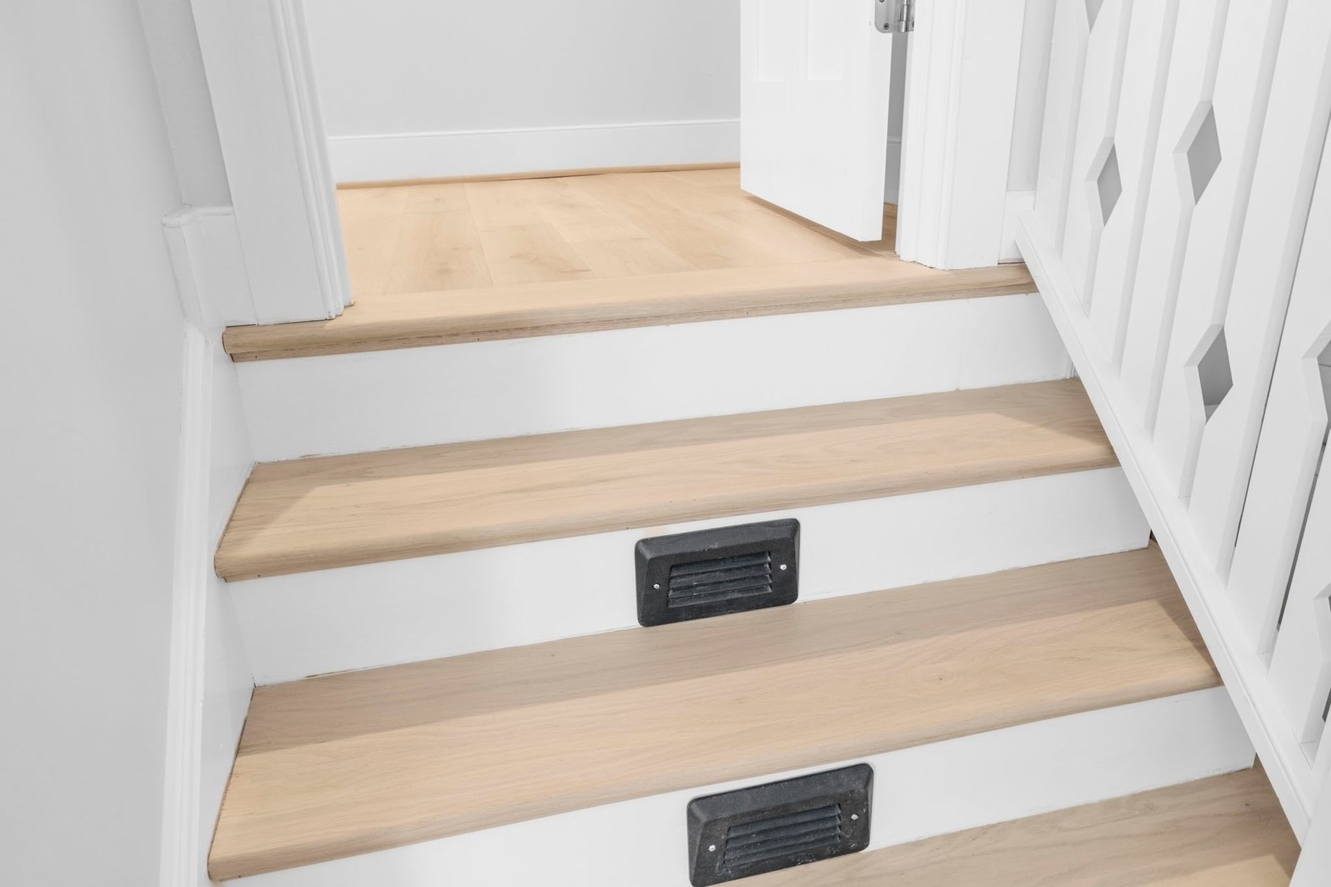 Indoor staircase with light wood treads, white walls, and black vent covers on the risers