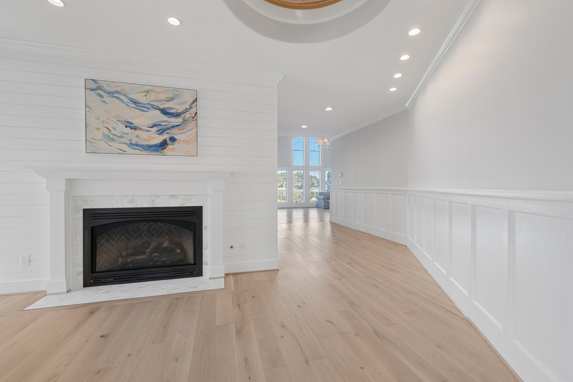 Bright white living room with fireplace, light wood floors, and a long hallway leading to windows.