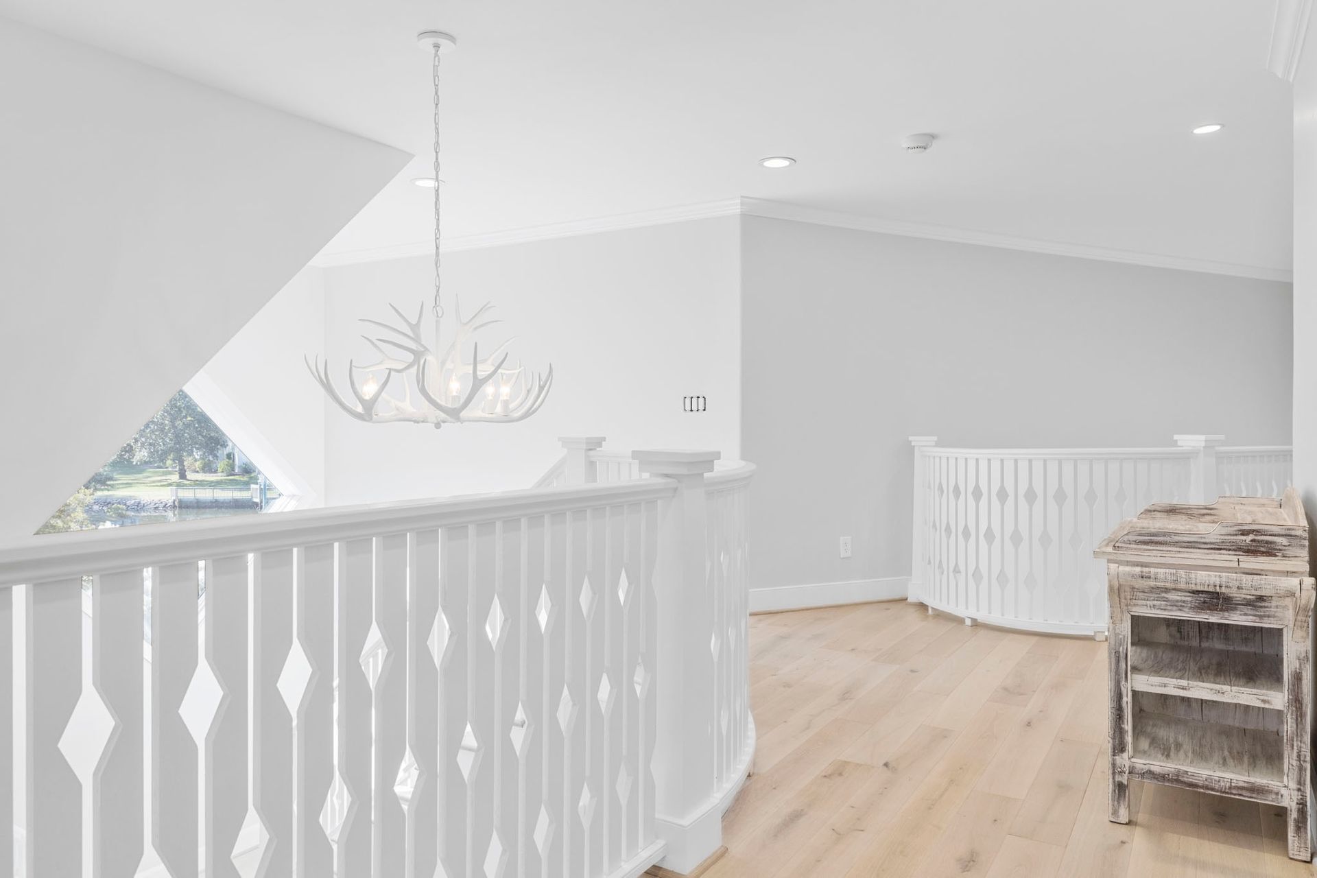 Bright upstairs hallway with white railings, chandelier, and light wood flooring, plus a wicker table.