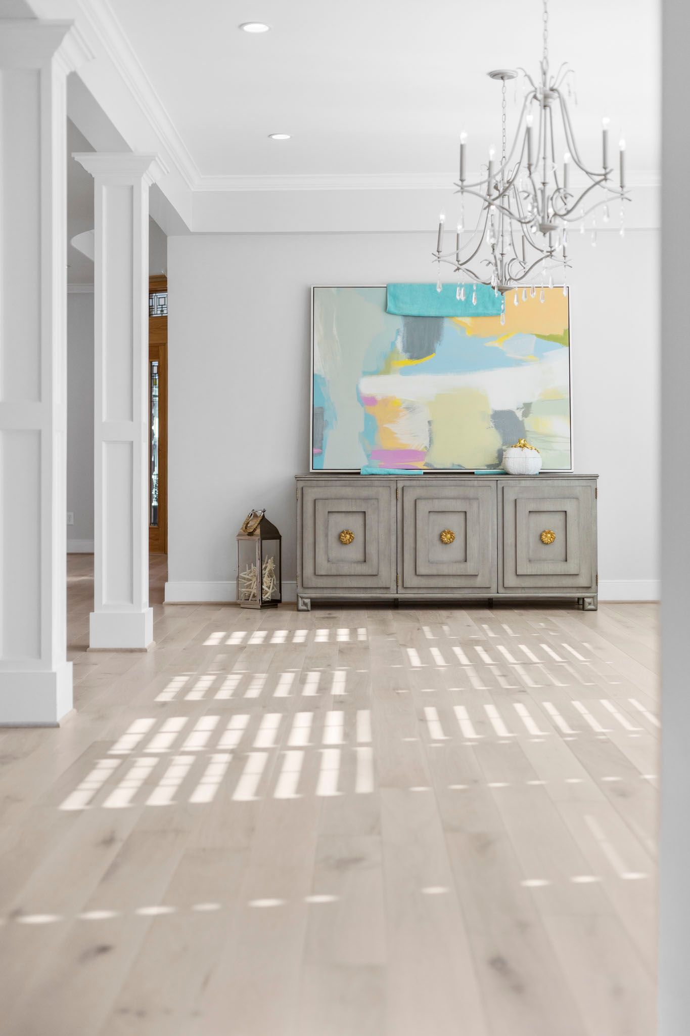 Bright hallway with white columns, chandelier, gray cabinet, colorful wall art, and sunlight on pale wood floors